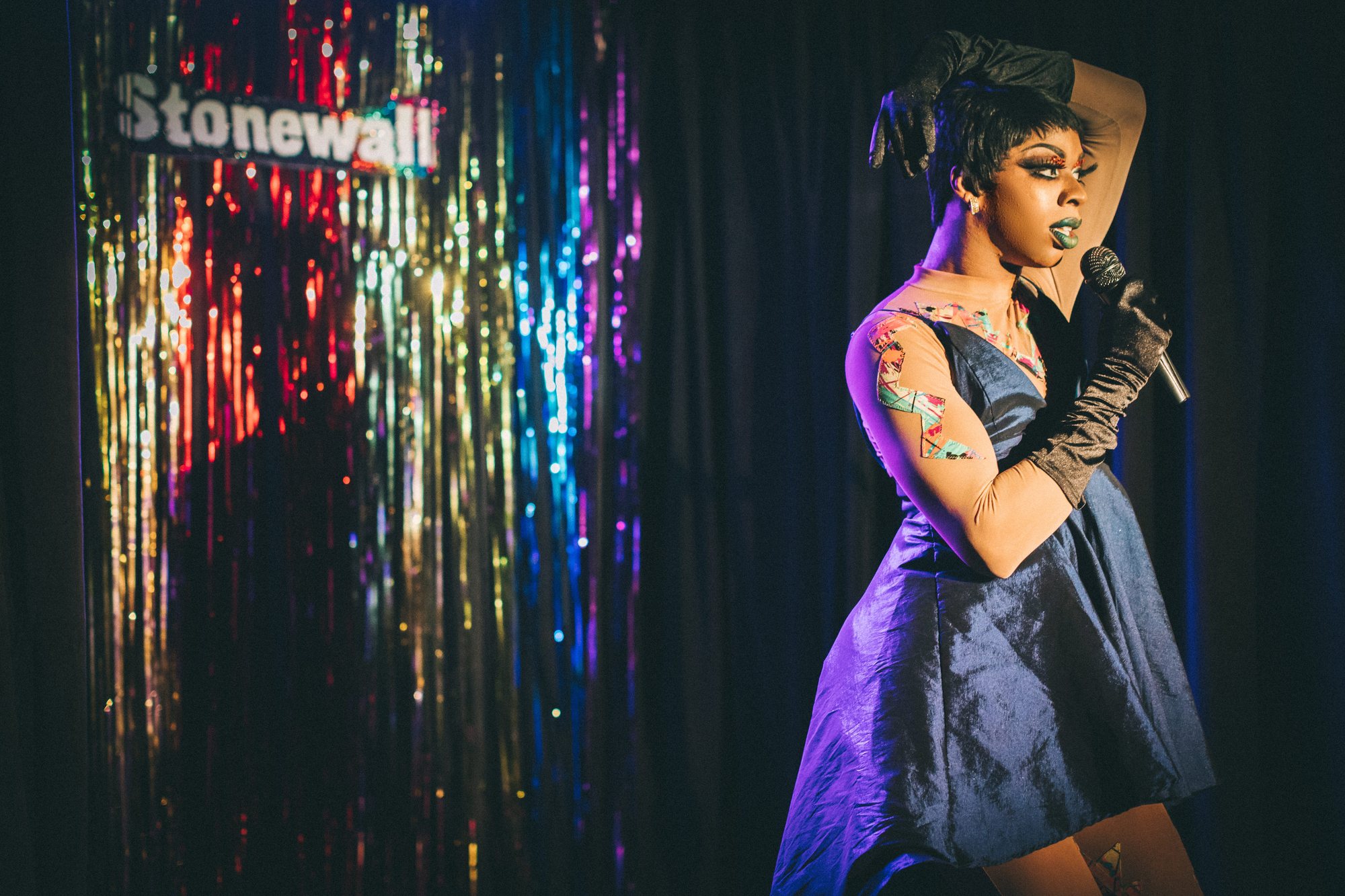 A drag performer singing in front of a rainbow tinsel curtain at Stonewall