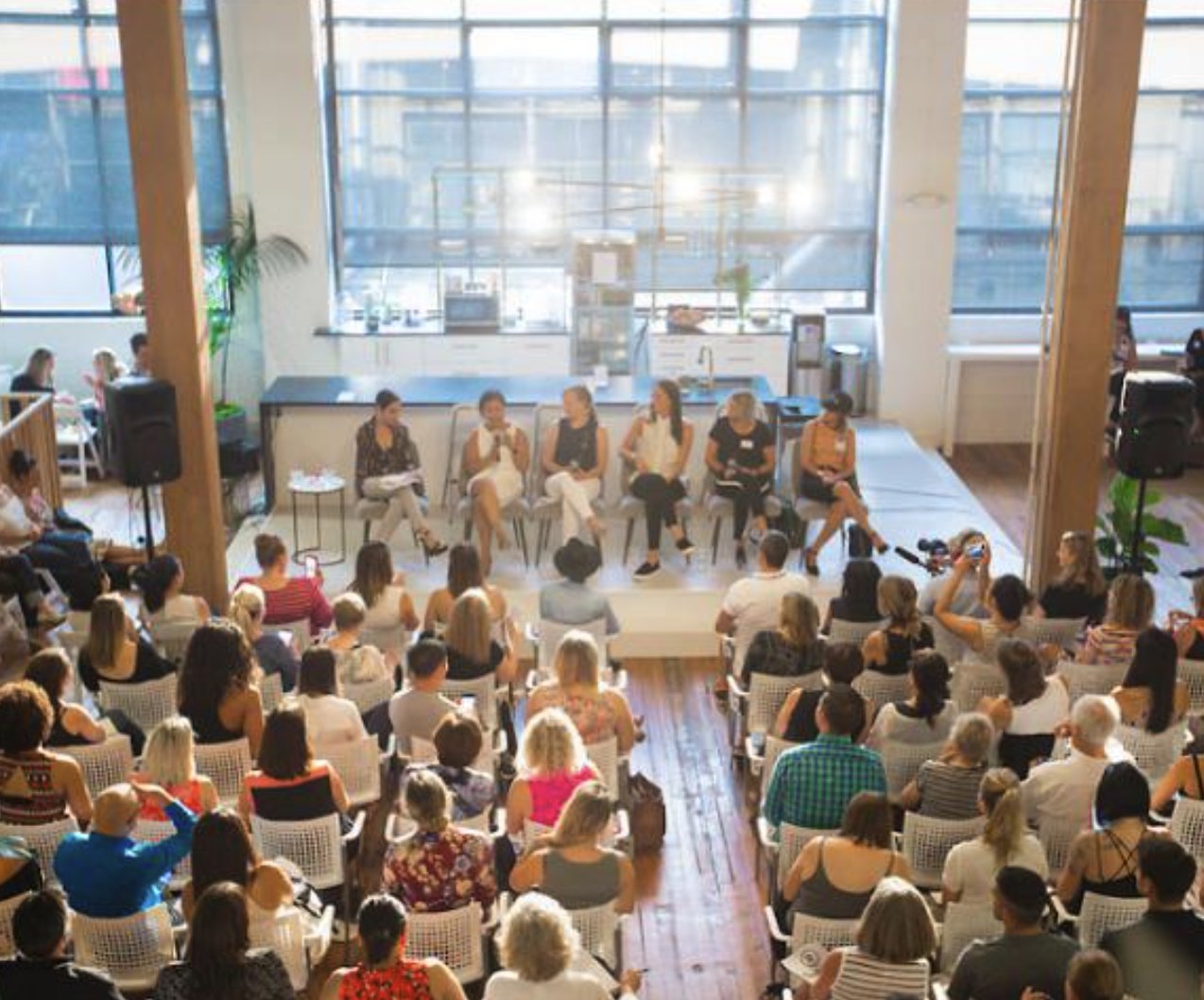 A panel of six on stage above a packed room of seated guests, tall windows behind