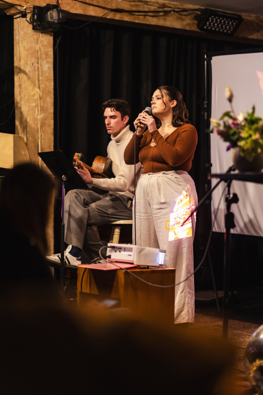 A singer at a microphone beside a seated guitarist on a warmly lit stage
