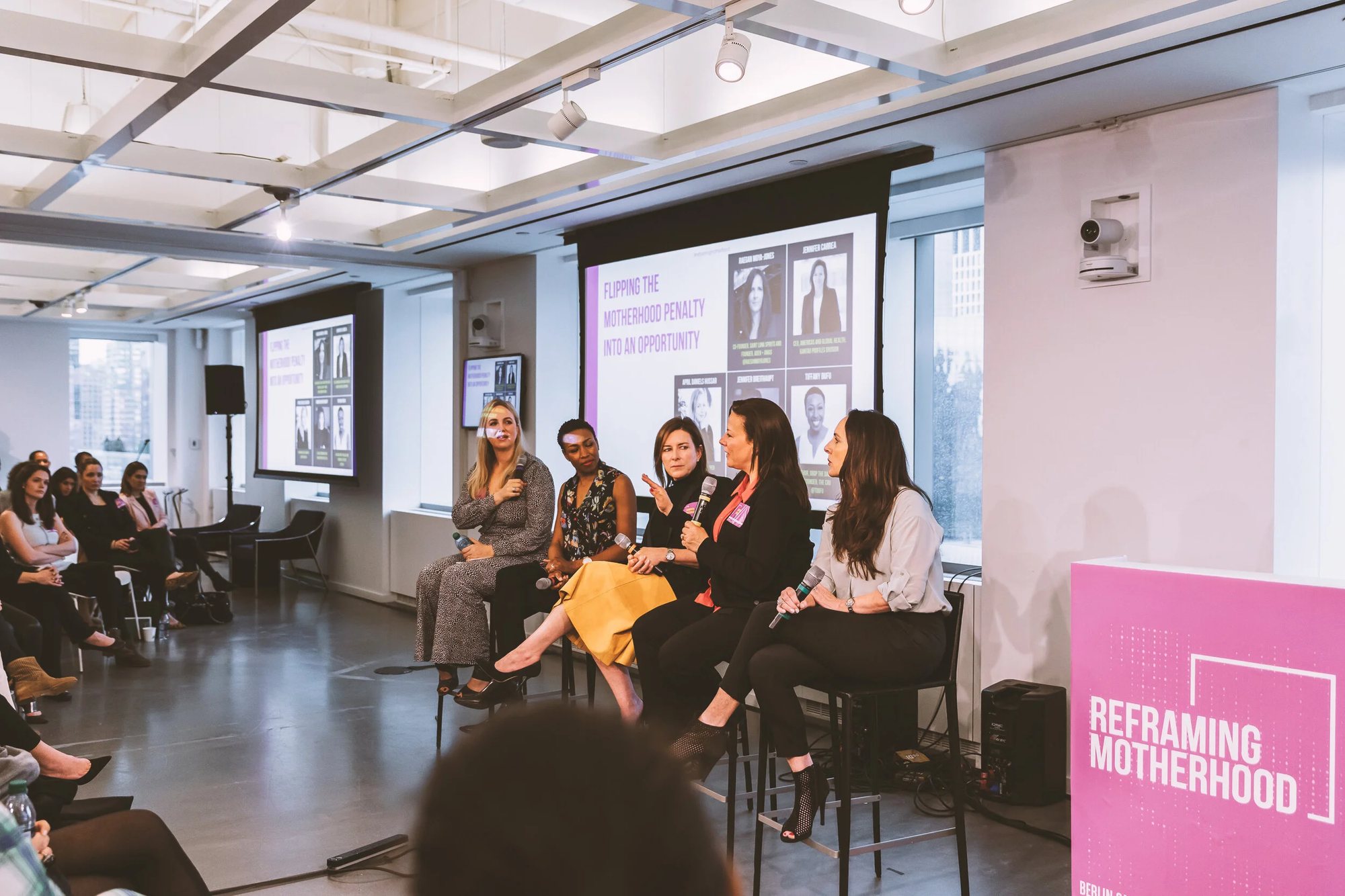 Five panellists on stage beside a pink Reframing Motherhood banner