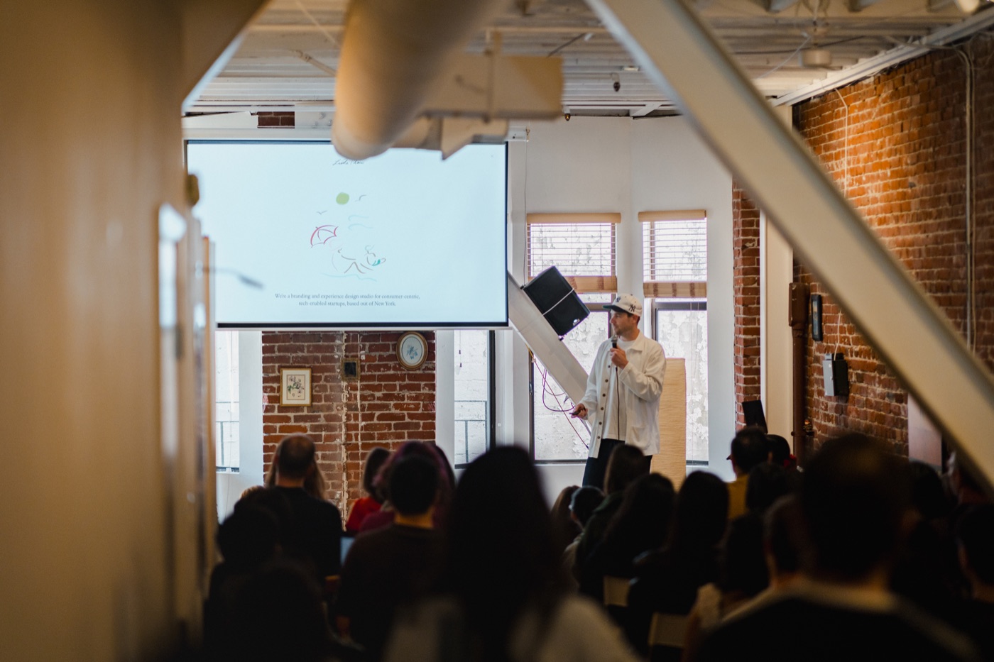 A speaker with a microphone presenting a slide to a seated audience in a brick loft