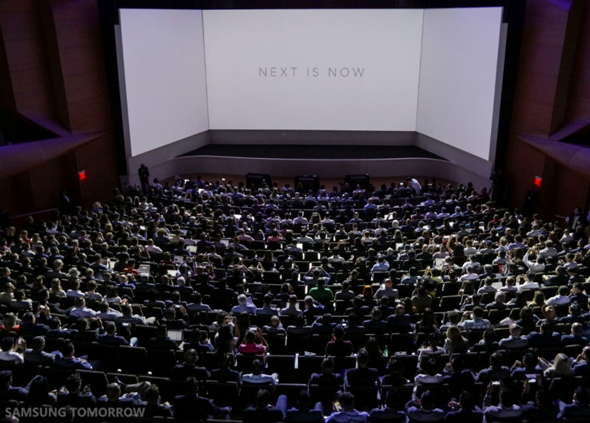 A packed auditorium facing a wraparound screen reading Next Is Now at Samsung Unpacked