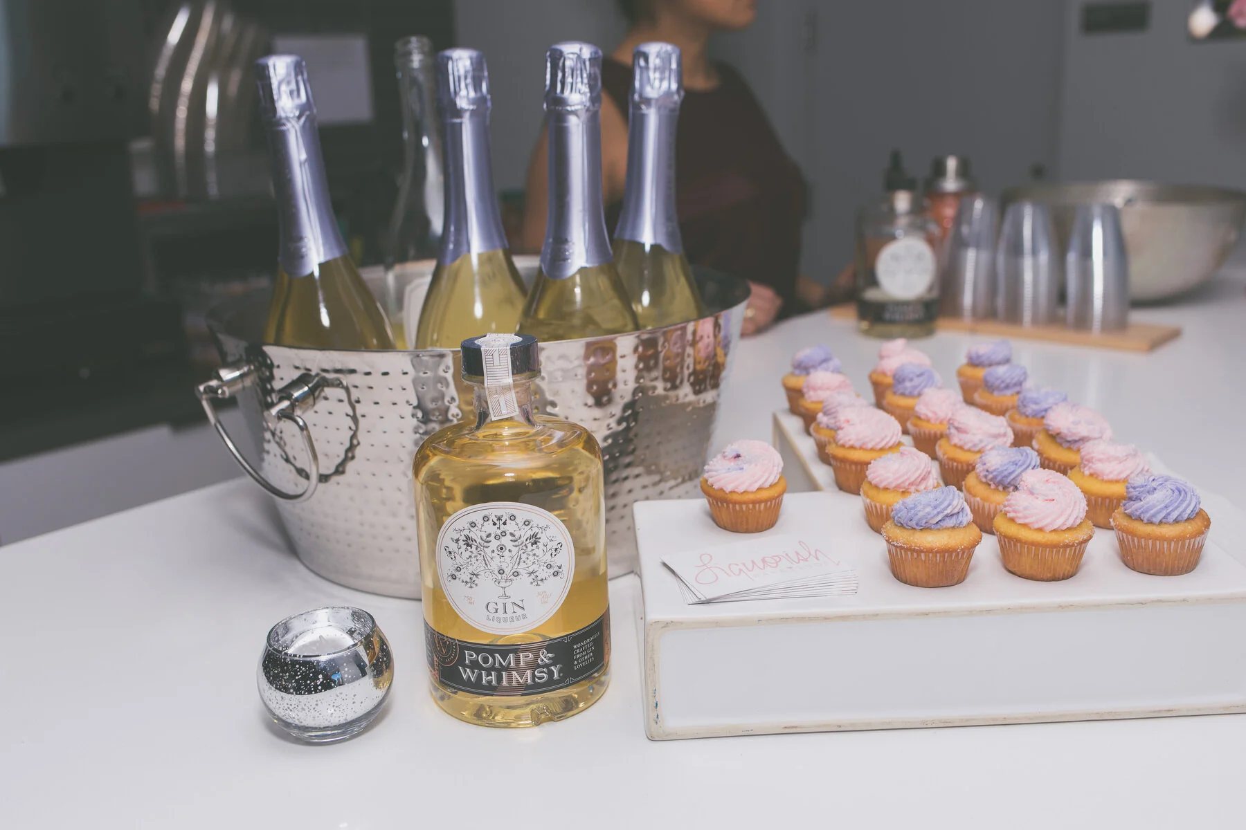 A bar table set with sparkling wine on ice, a bottle of gin and trays of pink and lilac cupcakes