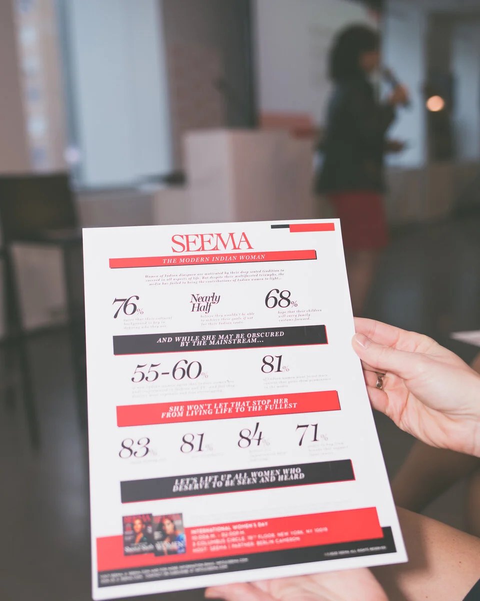 A hand holding the printed Seema programme, a speaker out of focus behind