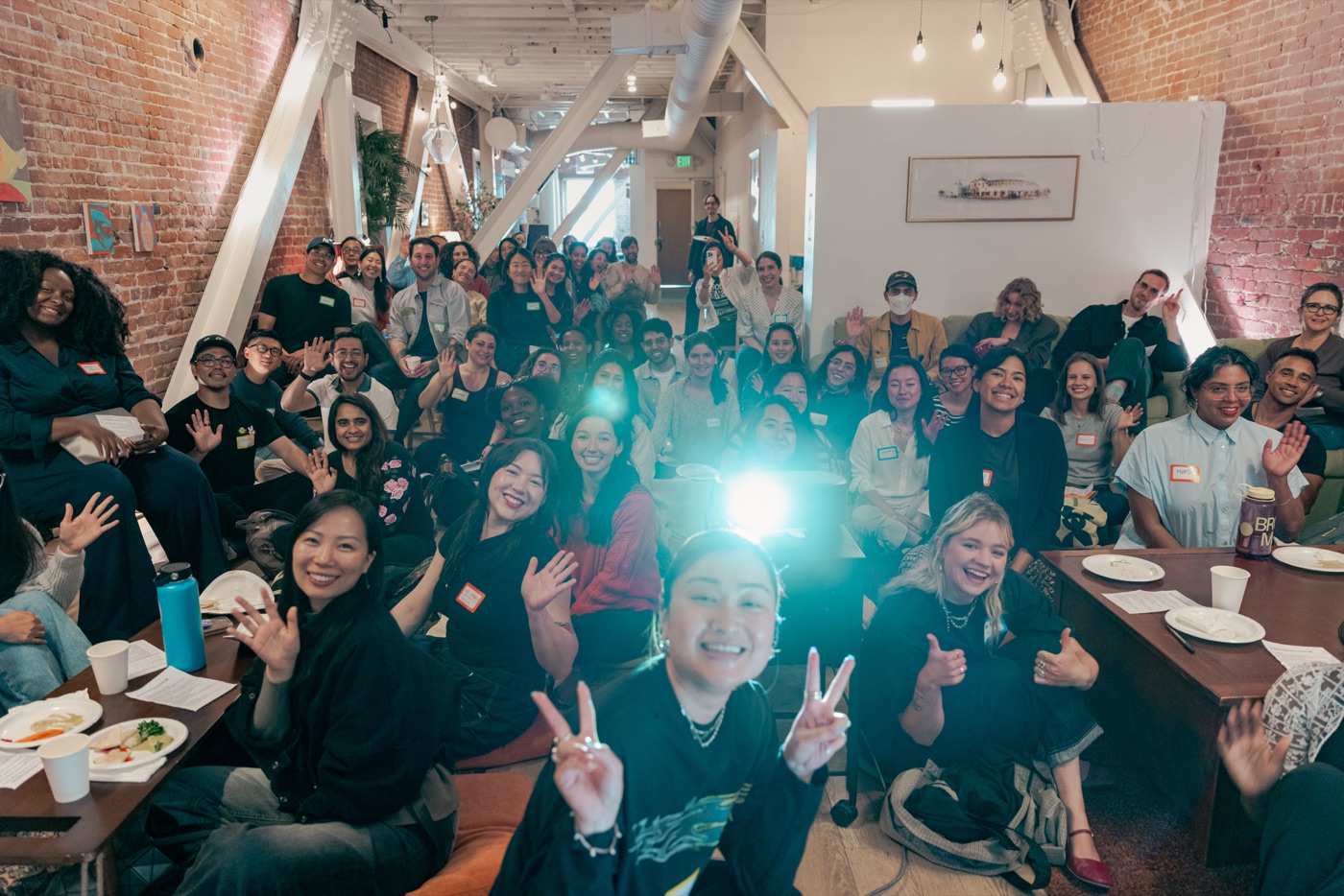 A packed room of guests seated on the floor, waving and smiling at the camera