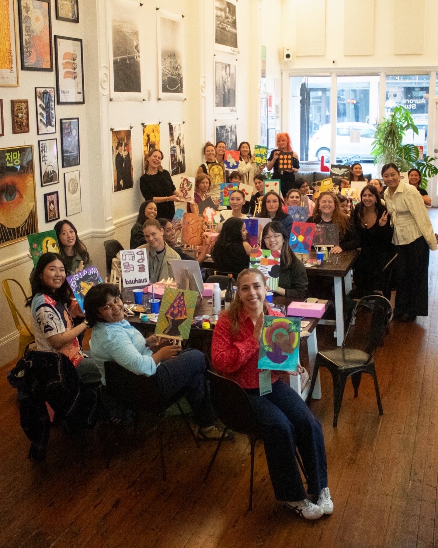 Guests holding up finished paintings around a table of pink Pour Toi boxes, in a gallery hung with prints